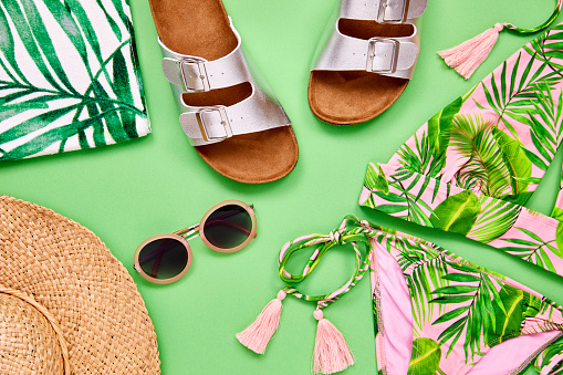 Flat lay shot of summer vacation accessories. Overhead shot of bikini sandal sunglasses and sun hat. They are arranged on green background.