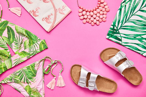 Flat lay color block shot of summer vacation accessories. Overhead shot of bikini sandal necklace and purse. They are arranged on pink background.