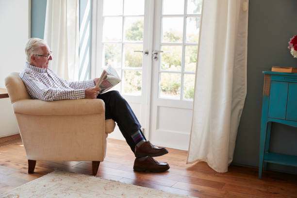 senior homme assis dans un fauteuil, lisant le journal à la maison - vieux fauteuil photos et images de collection