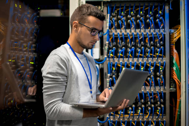 Man Managing Supercomputer Servers Side view  portrait of young man with laptop standing by server cabinet while working with supercomputer network connection plug stock pictures, royalty-free photos & images