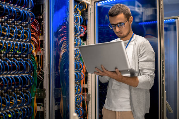 Supercomputer Manager Portrait of modern young man holding laptop standing in server room working with supercomputer in blue light computer connector stock pictures, royalty-free photos & images