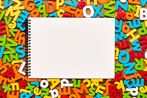 Overhead view of spiral notepad surrounded with multi colored alphabets on wood. Flat lay of blank notebook amidst letter magnets on wooden background. Empty space on book is for advertisement.