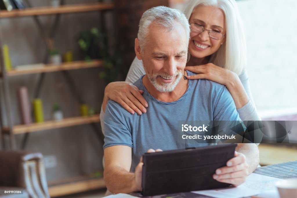 Retired positive man using tablet with his wife Happy family days. Waist up of a cheerful retired man using tablet and sitting at the table while resting together with his loving wife at home Baby Boomer Stock Photo