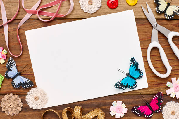 Overhead view of blank paper surrounded with craft products on wooden table. Artificial butterflies and art equipment are on plank. Empty space on can be used for advertisement purpose.