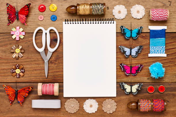 Overhead view of spiral notebook surrounded with craft products on wooden table. Artificial butterflies and decoration items are placed on wood. Blank space on paper can be used for advertisement.