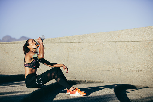 Healthy female sitting outdoors and drinking water after exercising session. Fitness woman taking a break after workout.