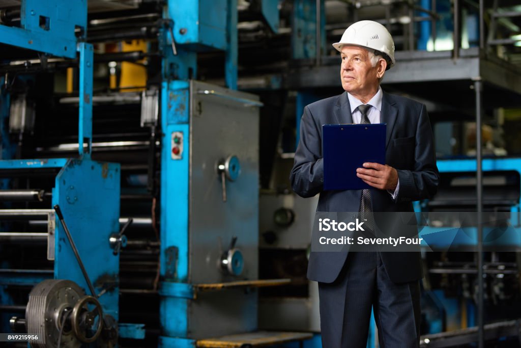 Confident Investor at Modern Plant Senior investor in suit and protective helmet taking necessary notes in clipboard while walking along production department of modern plant, portrait shot Hardhat Stock Photo