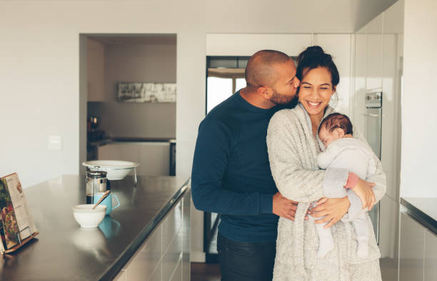 Lovely young family of three in kitchen Man kissing his wife holding a newborn baby boy in kitchen. Lovely young family of three in morning in kitchen. biracial newborn stock pictures, royalty-free photos & images