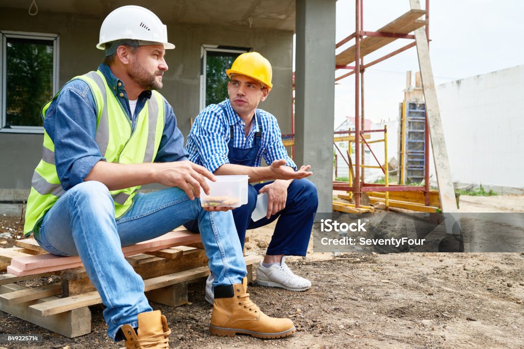 Taking Break from Work Two workers wearing protective helmets taking break from work and enjoying lunch while sitting outdoors, unfinished building on background Construction Worker Stock Photo