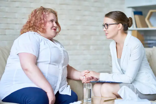 Obese young woman sharing problems with her highly professional psychologist, she holding hands and comforting her during therapy session, profile view