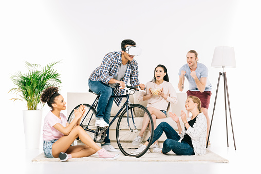 cheerful young multiethnic friends looking at young man in virtual reality headset riding bicycle