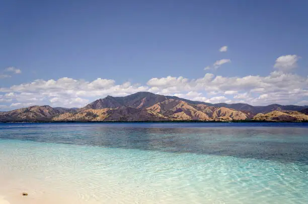 Empty white sand beach at midday overlooking broad island with green vegetation in te ocean, Flores Indonesia.