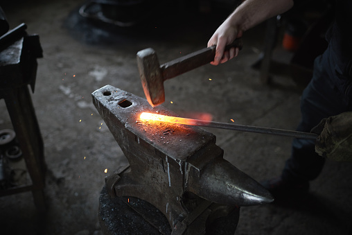 Close-up of unrecognizable person blacksmith bending wrought iron while hitting metal with hammer on anvil