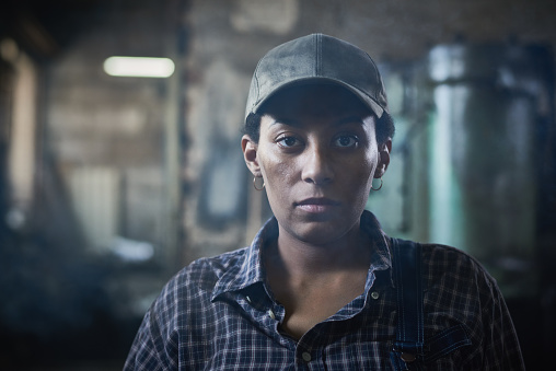 Unhappy young African-American female blacksmith in cap looking at camera in workshop