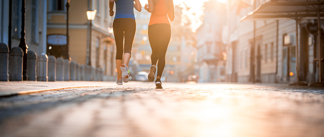 Two young women are running outdoors in the city on a summer day. They are wearing sports clothing and are focused on running.
