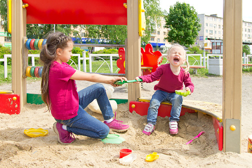 Conflict on the playground. Two sisters fighting over a toy in the sandbox. Kid sister crying all throat