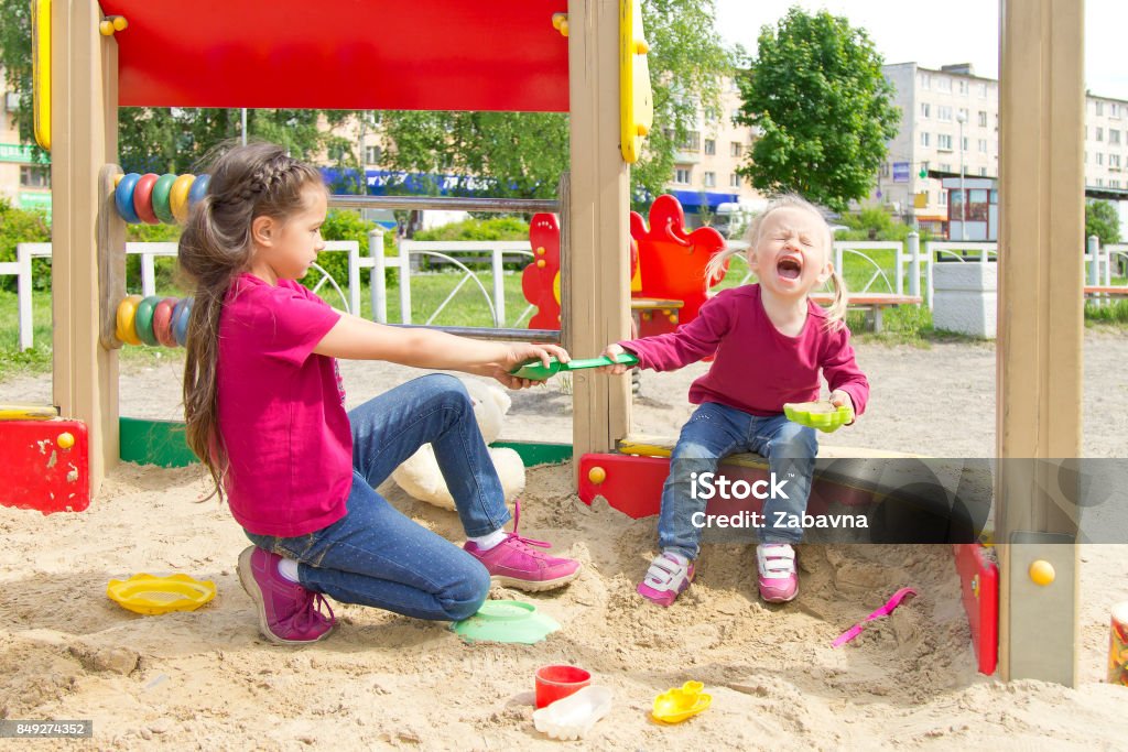 Conflict on the playground. Two sisters fighting over a toy in the sandbox. Kid sister crying all throat Conflict on the playground. Two sisters fighting over a toy shovel in the sandbox. Kid sister crying all throat Child Stock Photo