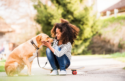 Beautiful african american girl with curly hair outdoors with her cute dog, sitting on skateboard.