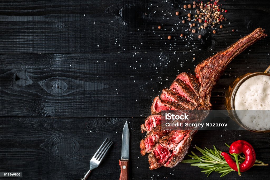 Barbecue dry aged rib of beef with spice, vegetables and a glass of light beer close-up on black wooden background Barbecue dry aged rib of beef with spice, vegetables and a glass of light beer close-up on black wooden background. Top view. Copy space. Still life. Flat lay Roasted Prime Rib Stock Photo