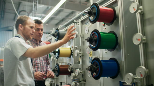Male engineers watching multicolor fiber optics spools on factory machine
