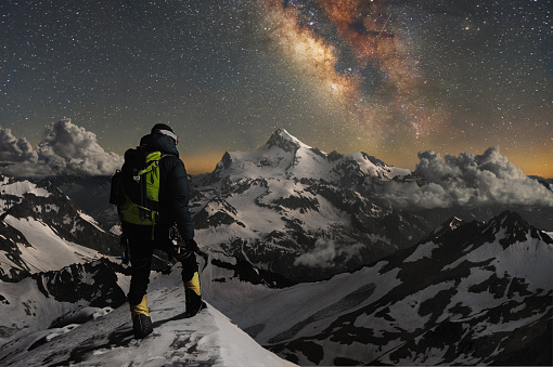Night photo. Professional mountaineering guide fully equipped and with a backpack on his shoulders and an ice ax in his hands stands on top of the mountain in the snow and looks at the surrounding mountains over which the starry sky and the milky way. Over the shoulders of the guide on the backpack hang carbines and folded sticks for Scandinavian walking. Long exposure.