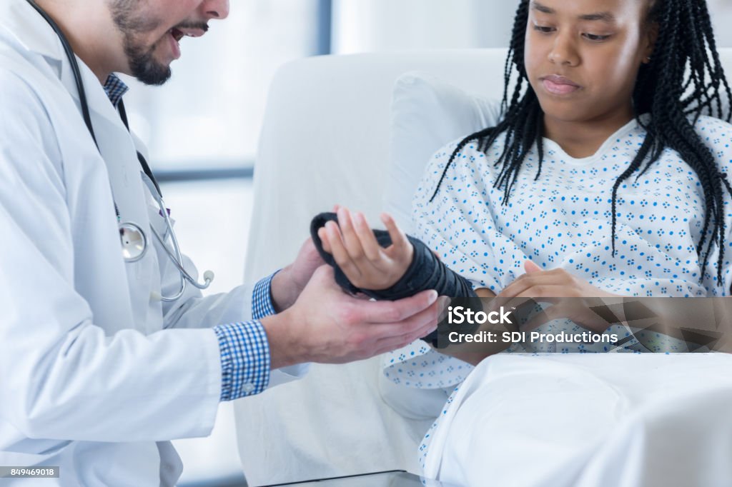 Teen hospital patient learns how to wear wrist brace A serious teenage girl wears a hospital gown and lays in a hospital bed as she holds her arm out to her unrecognizable physician.  Her doctor is showing her how to put on her new wrist brace. Emergency Medicine Stock Photo