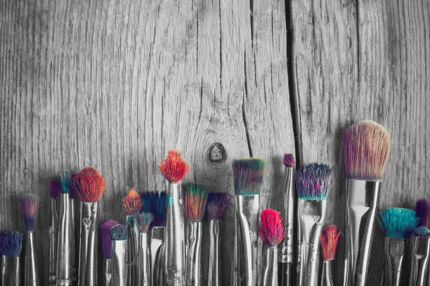 Row of artist paintbrushes with colorful bristle closeup on old wooden background, retro black and white stylized.