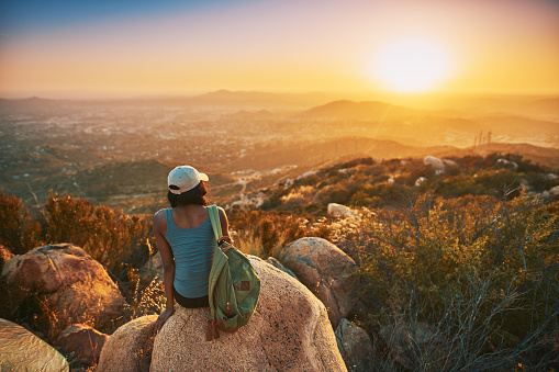 Rear view of woman hiker sitting on rock on top of hill while looking at sunset over San Diego California with colorful sky