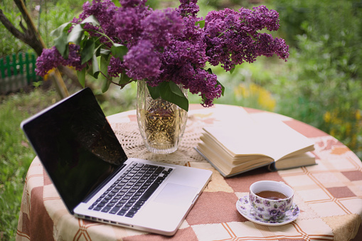 laptop, china teacup book and lilac bouquet in vase on green garden background