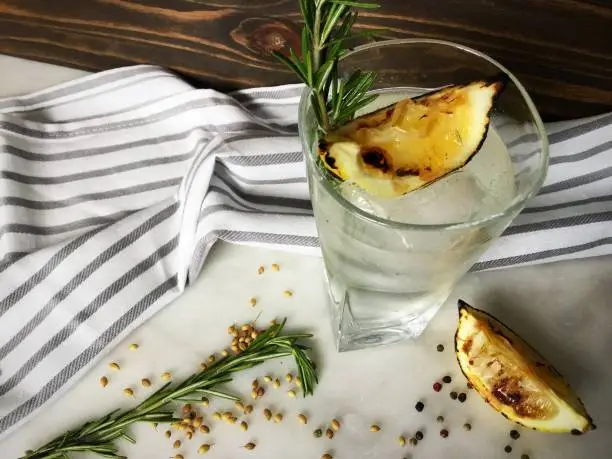A gin and tonic garnished with a rosemary sprig, charred lemon wedge and coriander seeds.