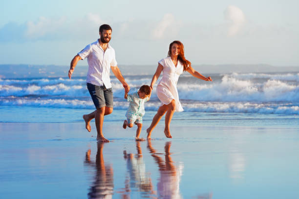 Family - father, mother, baby run on sunset beach Happy family - father, mother, baby son have fun together, child run with splashes by water pool along sunset sea surf on black sand beach. Travel lifestyle, parents with kids on summer vacation. raro stock pictures, royalty-free photos & images