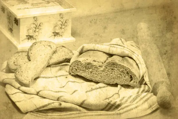 Homemade bread. Bread loaf wrapped in a kitchen towel, rolling pin next to it, and two slices of bread lying on the cloth. Decoupage box with roses is partially visible. Made to look like an old black-and-white photo. Selective focus.