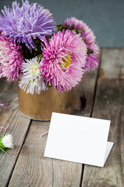 istock Multicolor aster flowers bouquet, blank white greeting card on rustic wooden table and dark grey background. Postcard mock up. Autumn flowers. Selective focus. Space for text. 849801154