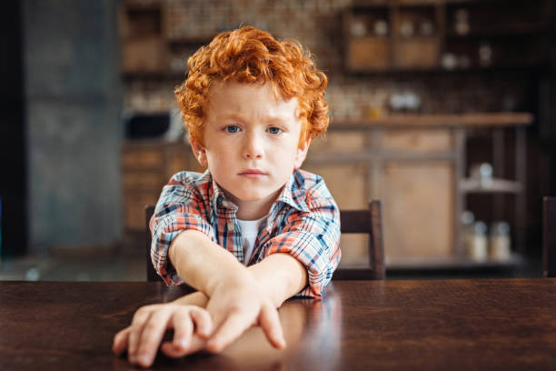 Bored redhead little boy sitting at table I am not in a playful mood today. Moody curly haired child sitting at a wooden table and stretching his hands while looking into the camera with eyes full of sadness. red head child sad stock pictures, royalty-free photos & images