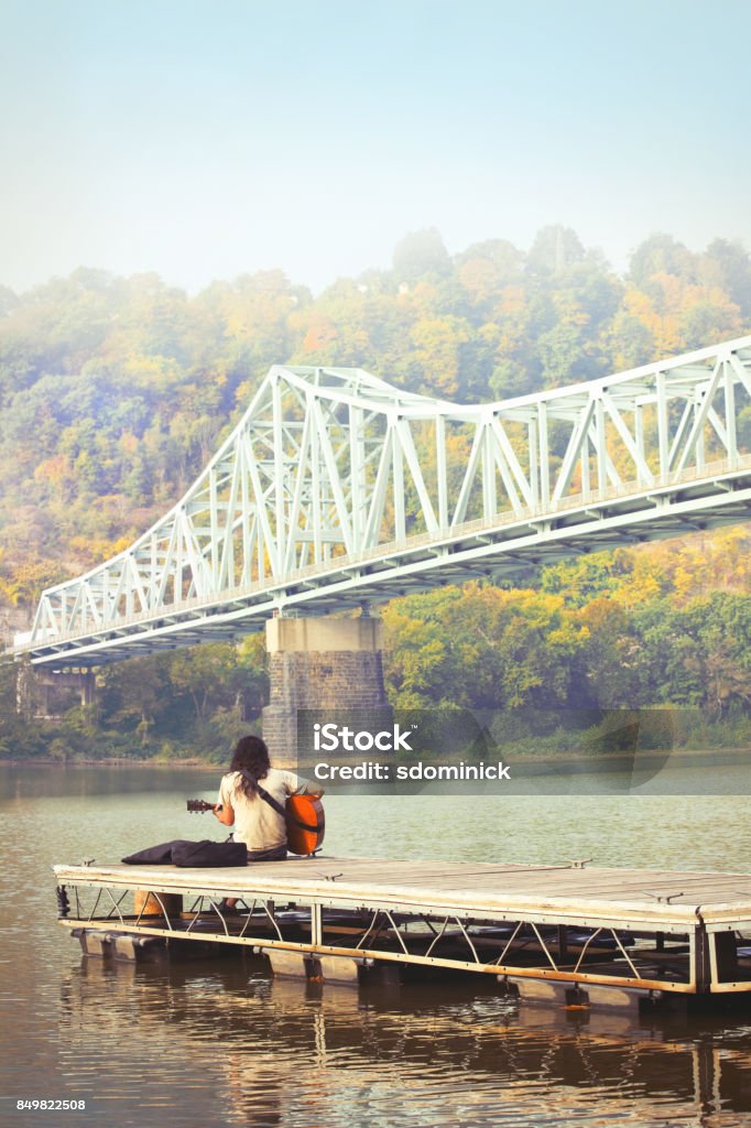 Man Playing Guitar On Boat Dock on Beautiful Fall Morning A man sitting on a boat dock playing his guitar in the morning on a beautiful fall day by the Ohio River in Pennsylvania by the Sewickley Bridge. Ohio River Stock Photo