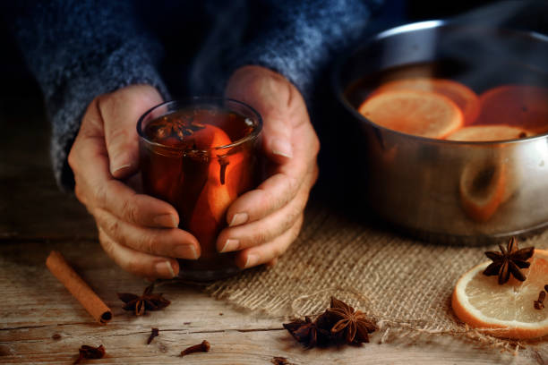 mani femminili più anziane che tengono una tazza di vetro con vin brulè caldo accanto alla pentola fumante, ingredienti natalizi, fette d'arancia, bastoncini di cannella, anice stellato e chiodi di garofano, una scena casalinga riscaldante su un tavolo d - drink brown herb heat foto e immagini stock