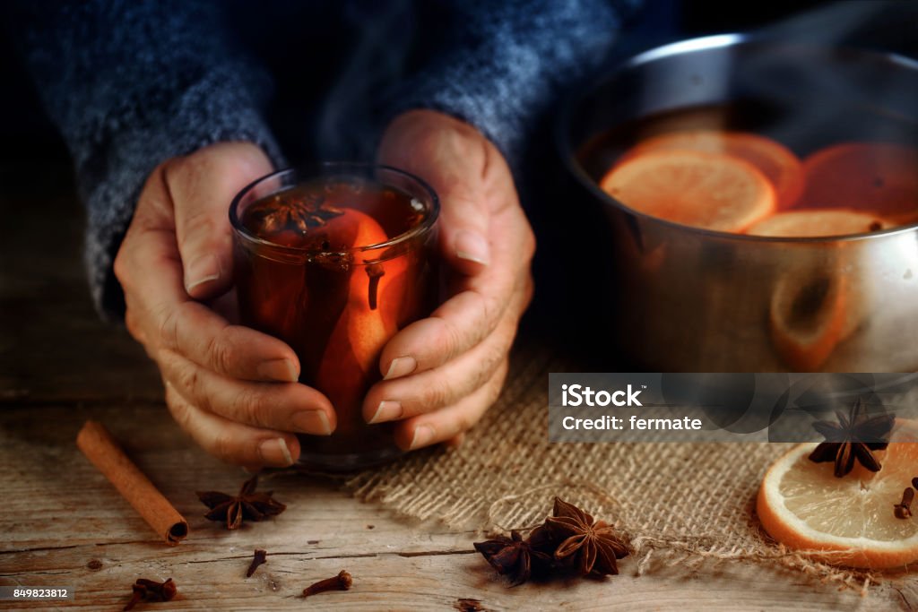 Older female hands holding a glass mug with hot mulled wine next to the steaming cooking pot,  Christmas ingredients, orange slices, cinnamon sticks, star anise and cloves, a warming home scene on a rustic wooden table Older female hands holding a glass mug with hot mulled wine next to the steaming cooking pot,  Christmas ingredients, orange slices, cinnamon sticks, star anise and cloves, a warming home scene on a rustic wooden table, selected focus, narrow depth of field Christmas Stock Photo