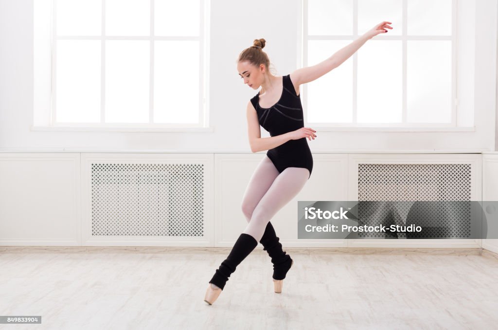 Beautiful ballerina stands in ballet assemble Young graceful ballerina in black at ballet class making assemble. Classical dancer in white hall practicing positions near large window, copy space Dancing Stock Photo