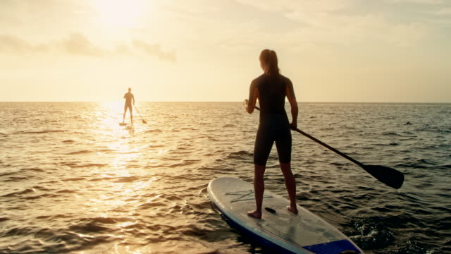 Woman standup paddling behind the man at sunset