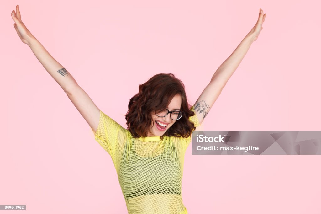 Excited hipster girl holding hands up Cheerful woman in trendy clothing and glasses looking happy with hands up on pink background. Colored Background Stock Photo