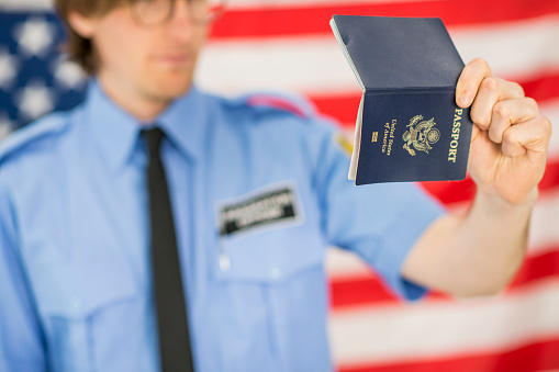 An immigration officer at a country's border is waiting to screen people through the border. He wears a uniform. Here he reads over a person's American passport while standing in front of the American flag.