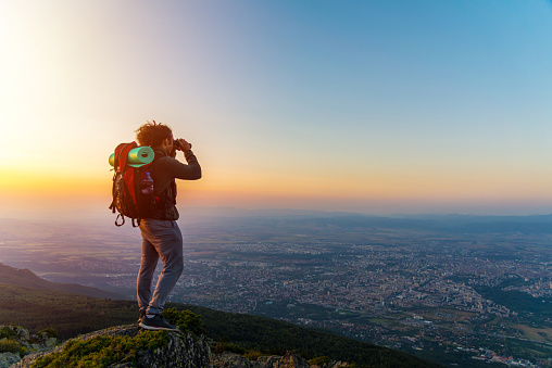 We see a guy who is watching the city through binoculars. He is hiking in the mountain and enjoys the view.