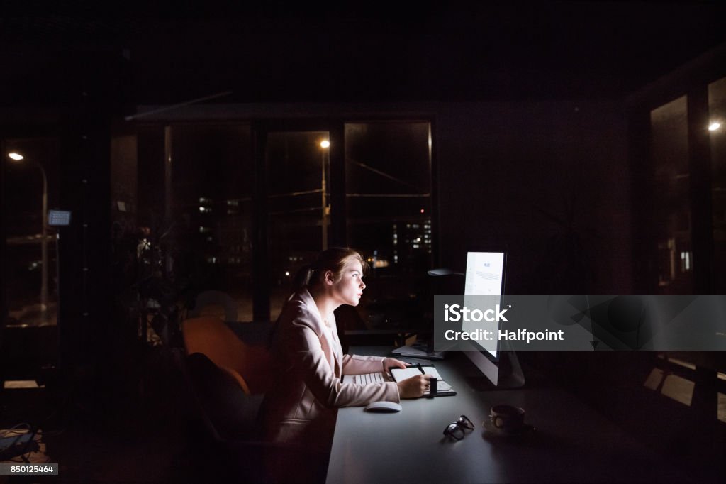 Businesswoman in front of computer screen in office at night. Beautiful young businesswoman in front of computer screen in her office late at night, writing into her personal organizer. Night Stock Photo