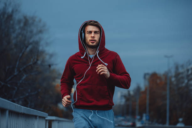 Hombre en rojo con capucha para correr al lado de la carretera en la ciudad - foto de stock