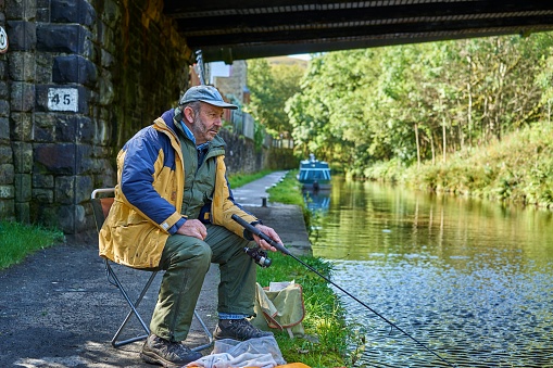 Senior man fishing with rod from the towpath of the Huddersfield Narrow Canal, Marsden, West Yorkshire, England. A distant canal boat is seen centre-frame beyond the bridge.