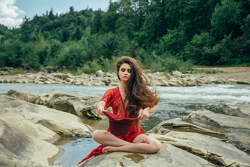 A nice girl sits on a rock in the middle of a boiling river and asks for alms from nature. Background wild nature. Creative colors.