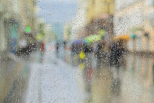 Raindrops on window glass, people walk on road in rainy day, blurred motion abstract background. View from the window on city street. Concept of shopping, modern city, walking, lifestyle