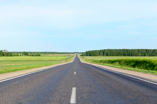 A small rural asphalt road. Landscape with blue sky, grass and trees. On the roadway cars are moving