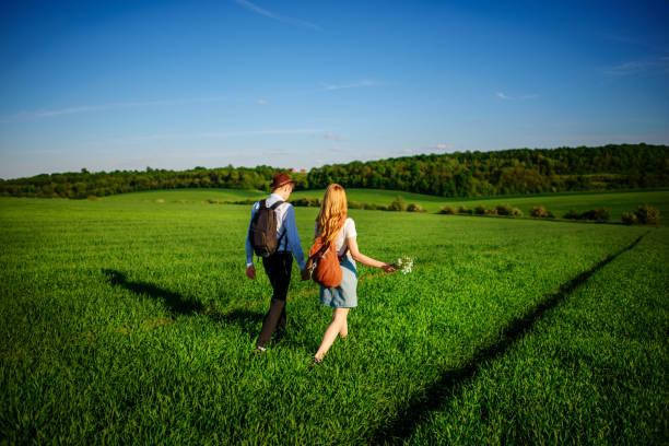 バックパック、帽子と長い髪を持つ女性で男はパスに沿って進みます。カップルは牧草地に沿って歩く - マサチューセッツ州 グリーンフィールド ストックフォトと画像