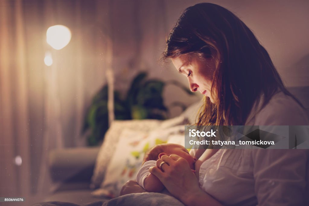 Young beautiful mother, breastfeeding her newborn baby boy at night Young beautiful mother, breastfeeding her newborn baby boy at night, dim light. Mom breastfeeding infant Breastfeeding Stock Photo